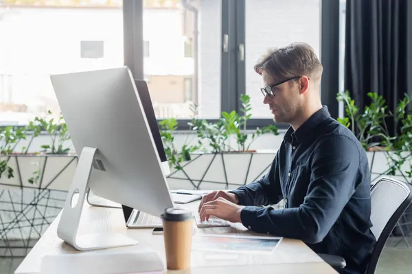 Seitenansicht des Programmierers mit Computern in der Nähe von verschwommenen Dokumenten und Kaffee im Büro — Stockfoto