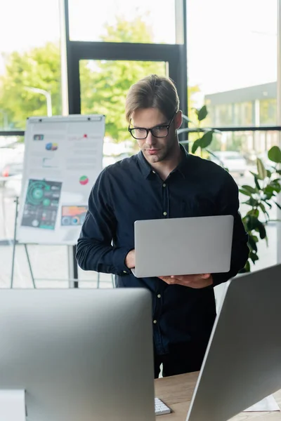 Giovane sviluppatore in occhiali da vista utilizzando laptop vicino a computer e lavagna a fogli mobili offuscata in ufficio — Foto stock