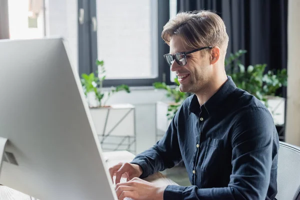 Smiling developer in eyeglasses using computer keyboard near monitor in office — Stock Photo
