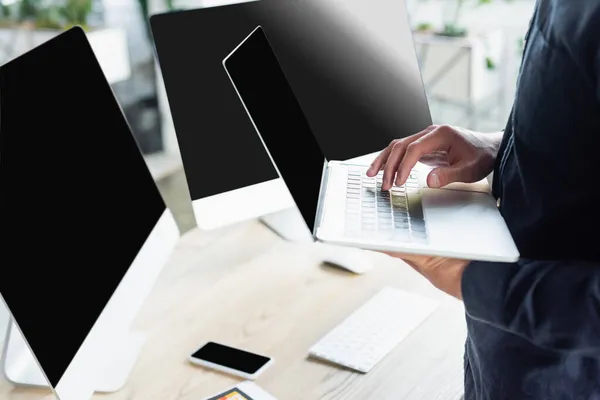 Vista recortada del programador usando el ordenador portátil cerca de computadoras con la pantalla en blanco en la oficina — Stock Photo
