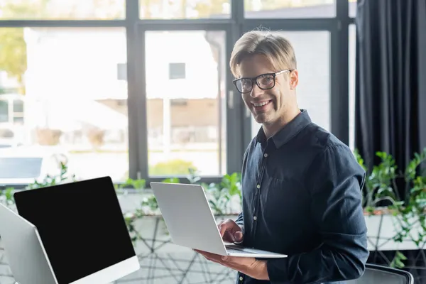 Молодой программист в очках улыбается в камеру при использовании ноутбука рядом с компьютерами в офисе — стоковое фото