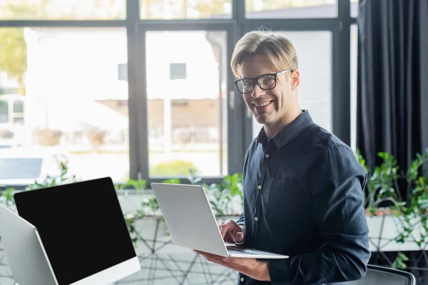 Joven programador en anteojos sonriendo a la cámara mientras usa el portátil cerca de computadoras en la oficina - foto de stock