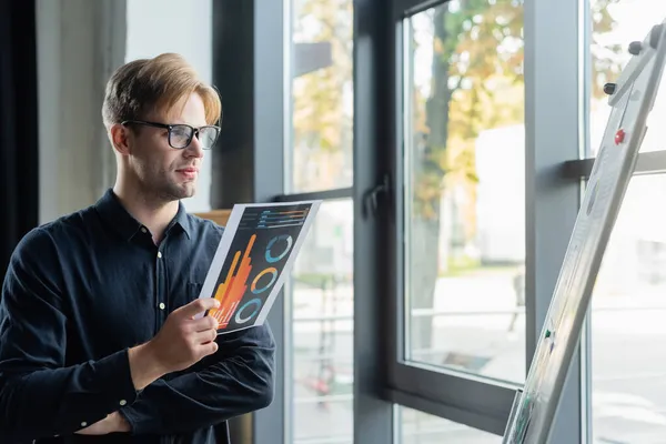 Joven programador en anteojos sosteniendo papel cerca de rotafolio y ventana en la oficina - foto de stock