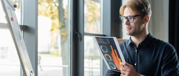 Developer in eyeglasses holding paper near flip chart in office, banner — Stock Photo