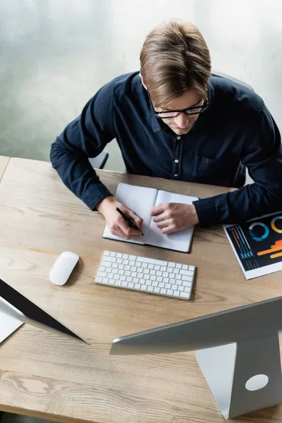 Overhead view of developer writing on notebook near computers in office — Stock Photo