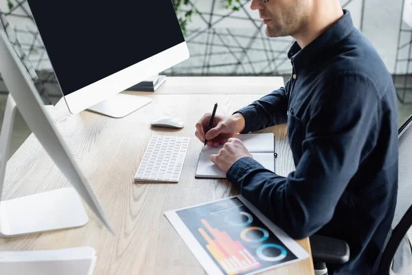 Cropped view of developer writing on notebook near computers and paper in office — Stock Photo