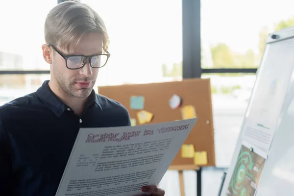 Young developer in eyeglasses holding blurred document near flip chart in office — Stock Photo