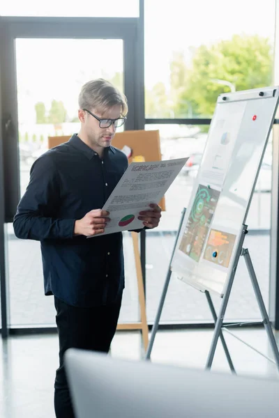 Programmatore in occhiali con documento vicino a lavagna a fogli mobili e computer sfocato in ufficio — Foto stock