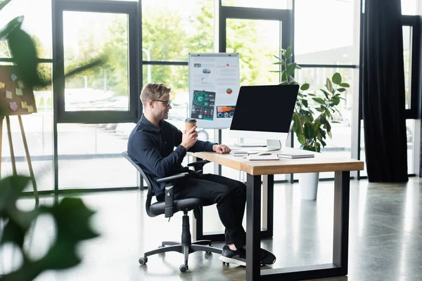 Seitenansicht eines lächelnden Entwicklers mit Kaffee in der Nähe des Computermonitors im Büro — Stockfoto