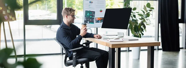 Vue latérale du programmeur souriant tenant boisson à emporter près de l'ordinateur avec écran blanc dans le bureau, bannière — Photo de stock