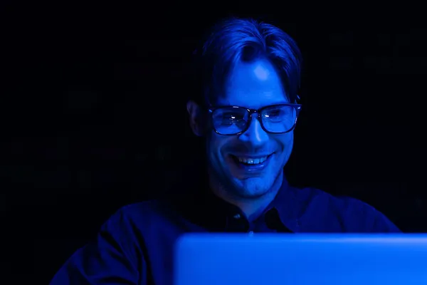 Smiling developer in eyeglasses looking at blurred computer monitor in office isolated on black — Stock Photo