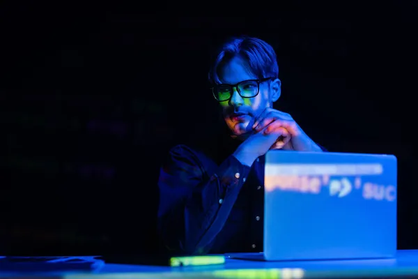 Sviluppatore in occhiali seduto vicino al computer con riflesso del codice in ufficio isolato su nero — Foto stock