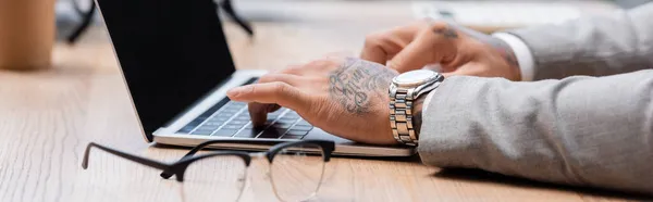Abgeschnittene Ansicht tätowierter Geschäftsmann in Armbanduhr tippt auf Laptop neben verschwommener Brille, Banner — Stockfoto