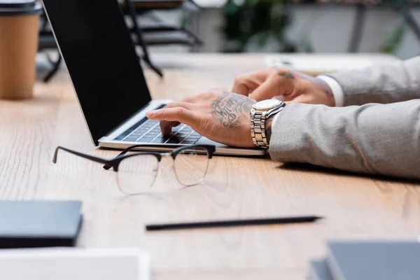 Vue partielle d'un homme d'affaires tatoué travaillant sur un ordinateur portable près de lunettes floues sur un bureau — Photo de stock