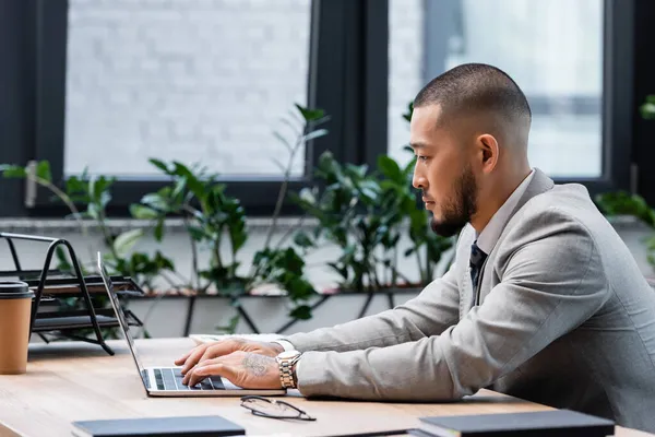 Seitenansicht eines bärtigen asiatischen Geschäftsmannes, der im Büro am Laptop arbeitet — Stockfoto