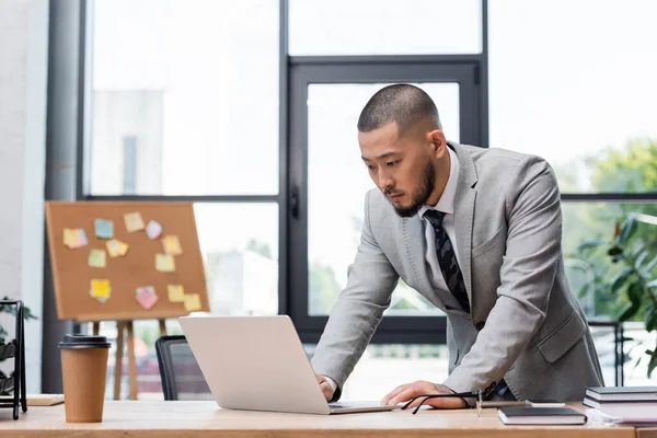 Barbudo asiático hombre de negocios de pie cerca de escritorio y mirando portátil en oficina - foto de stock