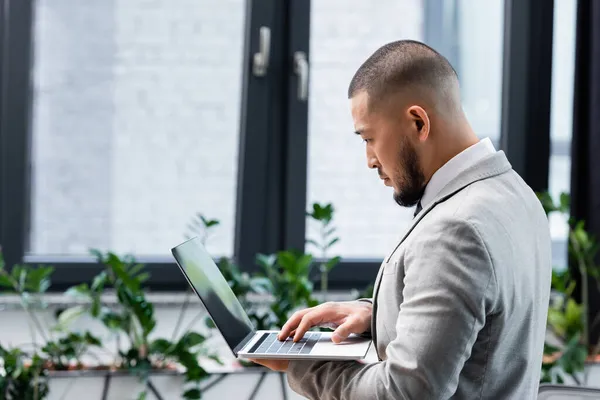 Asiático hombre de negocios mecanografía en portátil mientras de pie en oficina - foto de stock