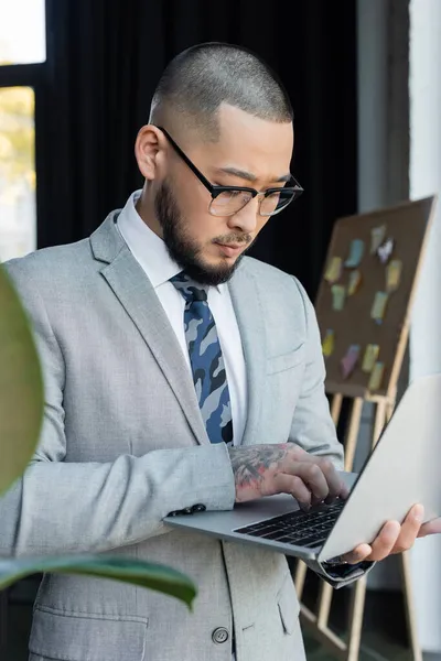 Tatuado ásia empresário no óculos usando laptop no embaçado primeiro plano no escritório — Fotografia de Stock