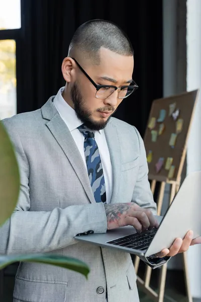 Tatuado asiático hombre de negocios en gafas usando portátil en borrosa primer plano en oficina - foto de stock