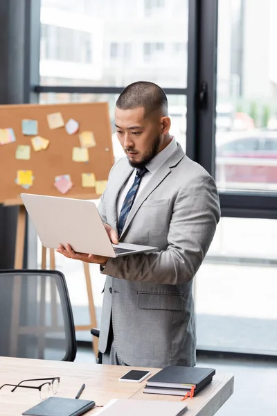 Asiático hombre de negocios en formal desgaste de pie con portátil cerca de escritorio en oficina - foto de stock