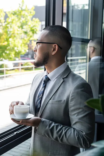 Asiático hombre de negocios con taza de café de pie cerca de ventana en oficina - foto de stock