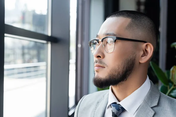 Bearded asian businessman in eyeglasses looking away near window in office — Stock Photo