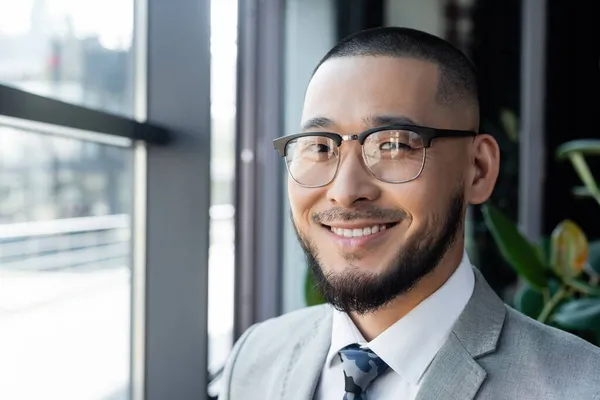 Barbuto asiatico uomo d'affari in occhiali sorridente a camera in ufficio — Foto stock