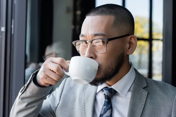 Asian businessman in formal wear and eyeglasses drinking coffee in office — Stock Photo