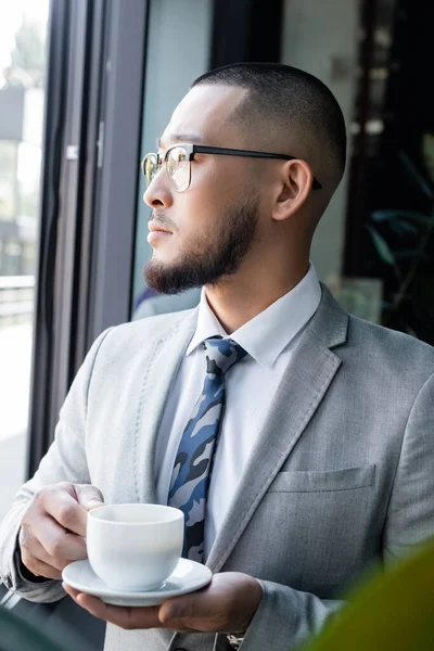 Asian businessman in formal wear and eyeglasses holding coffee cup in office — Stock Photo