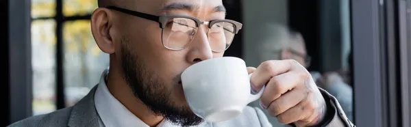 Close up view of asian businessman in eyeglasses drinking coffee in office, banner — Stock Photo