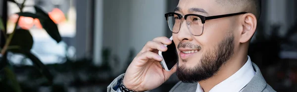 Позитивний азіатський бізнесмен в окулярах дивиться далеко під час дзвінка на мобільний телефон, банер — стокове фото