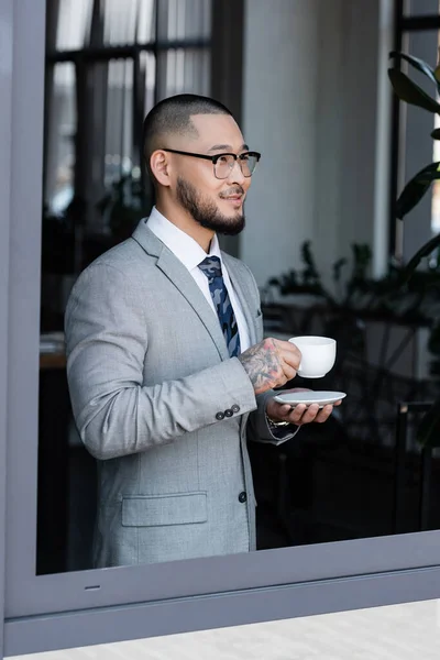 Sorridente uomo d'affari asiatico con una tazza di caffè in piedi vicino alla finestra in ufficio — Foto stock