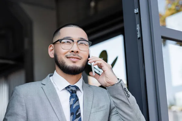 Glücklicher asiatischer Geschäftsmann in Brille, der am Fenster mit dem Handy spricht — Stockfoto