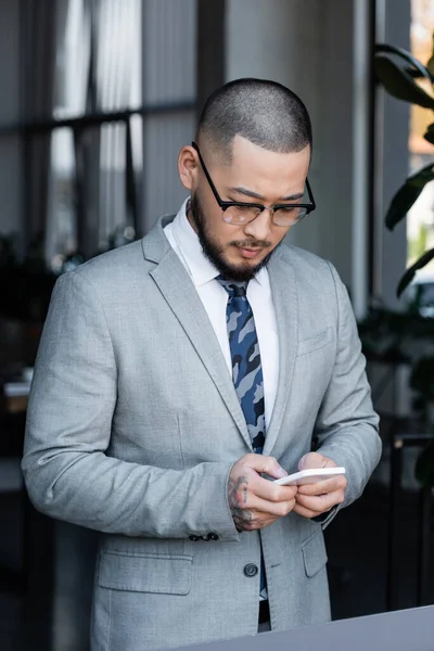 Asian businessman in formal wear and eyeglasses messaging on cellphone in office — Stock Photo