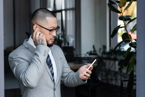 Tätowierter asiatischer Geschäftsmann setzt Kopfhörer ein, während er im Büro mit dem Handy telefoniert — Stockfoto