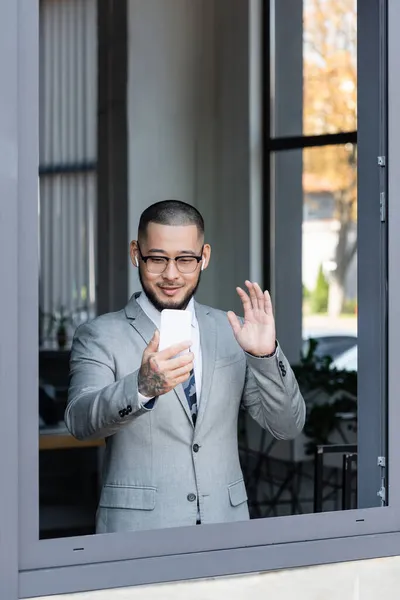 Улыбающийся азиатский бизнесмен в наушниках машет рукой во время видеочата на смартфоне возле окна офиса — стоковое фото