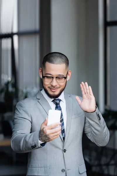 Sorridente asiático empresário em fones de ouvido acenando mão durante chamada de vídeo no smartphone — Fotografia de Stock