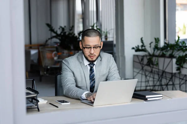 Asian businessman in eyeglasses working on laptop in office on blurred foreground — Stock Photo
