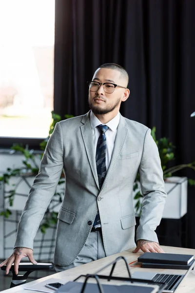 Asiático hombre de negocios en formal desgaste y gafas mirando hacia otro lado mientras de pie en escritorio - foto de stock