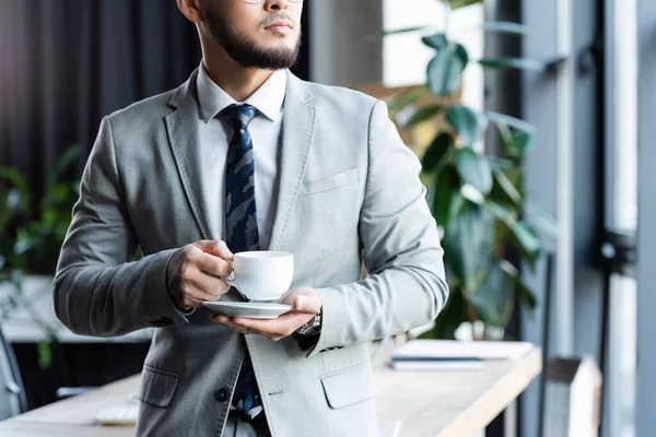 Cropped view of bearded businessman in formal wear standing with cup of coffee in office — Stock Photo