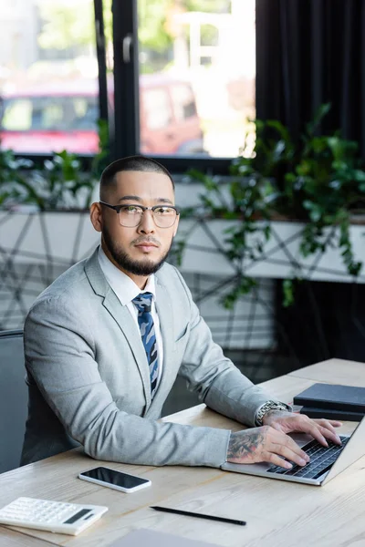 Asiatischer Geschäftsmann schaut auf Kamera, während er auf Laptop neben Smartphone mit leerem Bildschirm tippt — Stockfoto