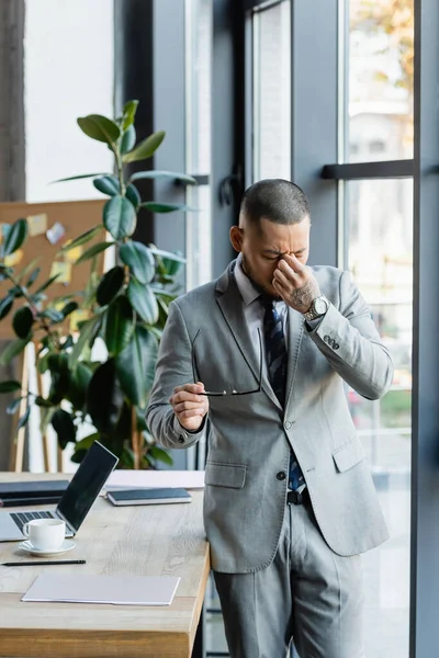 Tired asian businessman holding eyeglasses and rubbing eyes near work desk in office — Stock Photo