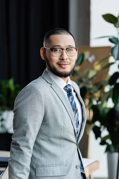 Счастливый азиатский бизнесмен в формальной одежде и очках, смотрящий в камеру в офисе — стоковое фото