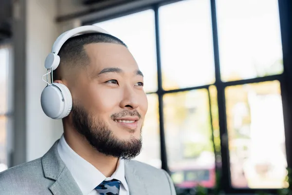 Feliz asiático homem de negócios em fones de ouvido sem fio olhando para longe no escritório — Fotografia de Stock