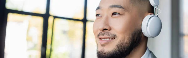Smiling asian businessman in wireless headphones looking away in office, banner — Stock Photo