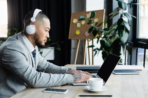 Asiático hombre de negocios en inalámbrico auriculares escribiendo en portátil con pantalla en blanco en la oficina - foto de stock