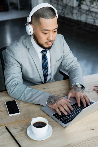 Азиатский бизнесмен в наушниках печатает на ноутбуке возле чашки кофе и смартфона с чистым экраном — стоковое фото