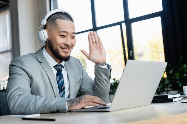 Smiling asian businessman in headphones waving hand during video call on laptop in office — Stock Photo
