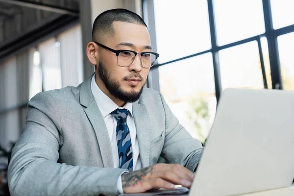 Asian businessman in eyeglasses typing on blurred laptop in office — Stock Photo