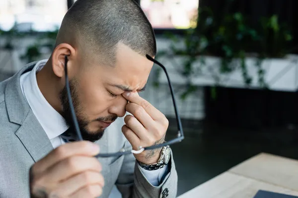 Tired asian businessman holding blurred eyeglasses while rubbing eyes in office — Stock Photo
